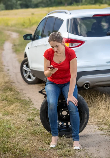 Mujer molesta sentada en un neumático de repuesto junto a un coche roto en el campo y —  Fotos de Stock