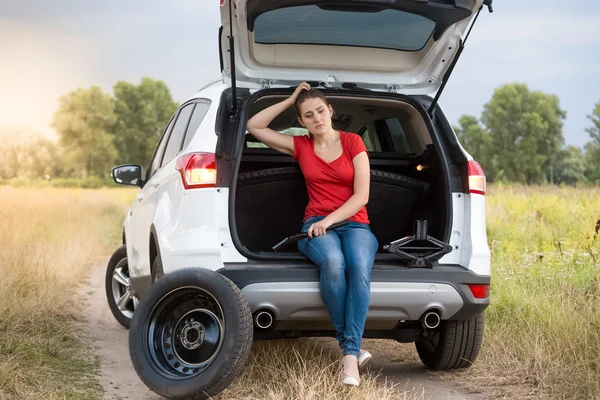 Sad woman sitting in open trunk of broken car on the rural road — Stock Photo, Image