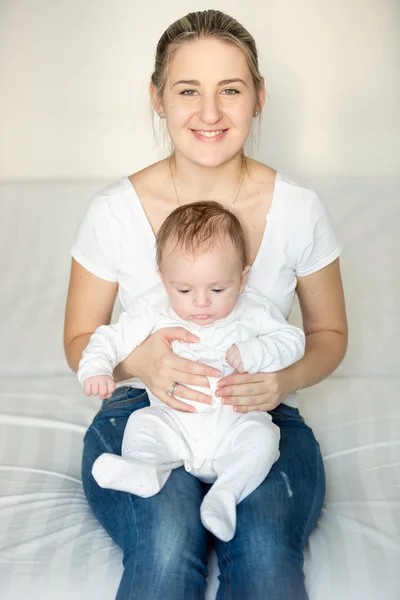 Mujer sonriente sentada en el dormitorio y sosteniendo a su lindo bebé en la — Foto de Stock