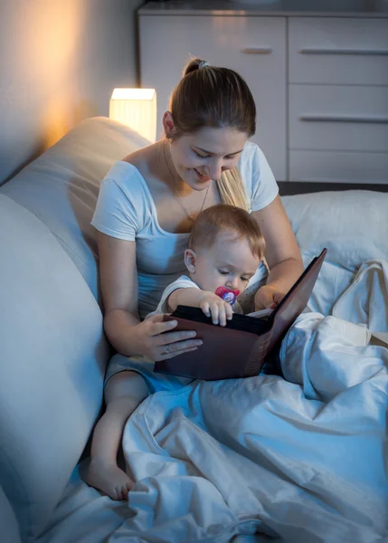 Hermosa madre joven leyendo libro a su bebé en la cama por la noche — Foto de Stock