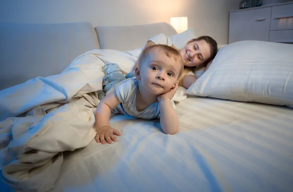 Funny portrait of baby boy lying on bed next to his mother tryin