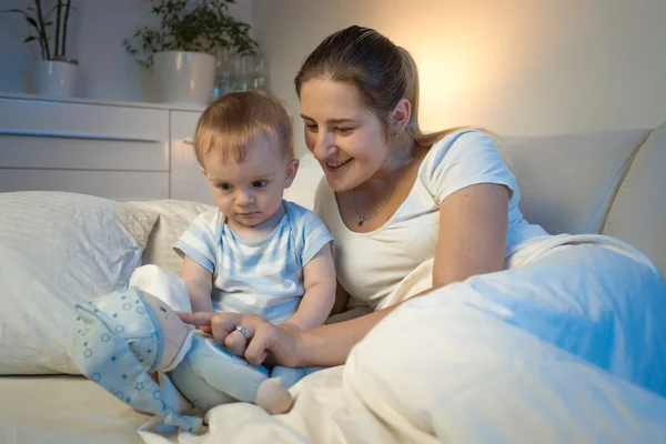 Lachende moeder spelen met haar baby liggend in bed bij nacht en p — Stockfoto