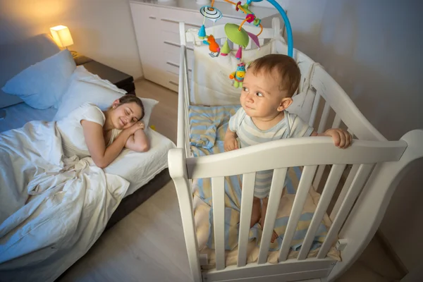 Retrato de bebé de pie en cuna y mirando a la madre cansada th — Foto de Stock