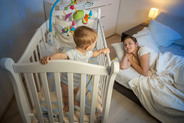 9 meses de edad bebé de pie en la cuna y despertar a su mothe cansado —  Fotos de Stock