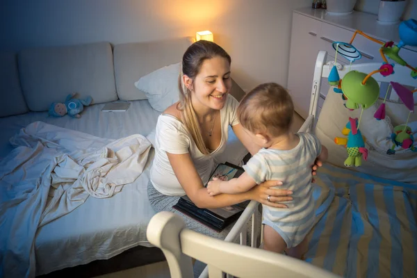 Portrait of beautiful mother taking baby out of crib at night — Stock Photo, Image