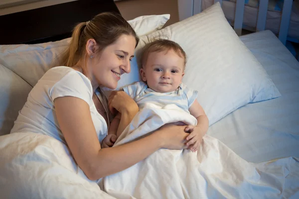 Hermosa mujer sonriente acostada en la cama por la noche y abrazando a su ba —  Fotos de Stock