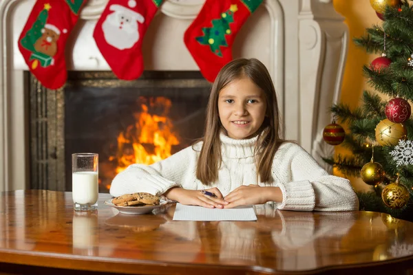 Feliz chica sonriente sentada junto a la chimenea y escribiendo una carta a Sa —  Fotos de Stock