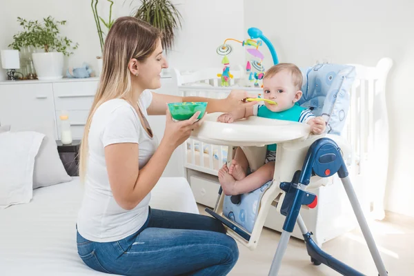 Bela mãe alimentando seu bebê menino em cadeira alta com mingau de cereal — Fotografia de Stock