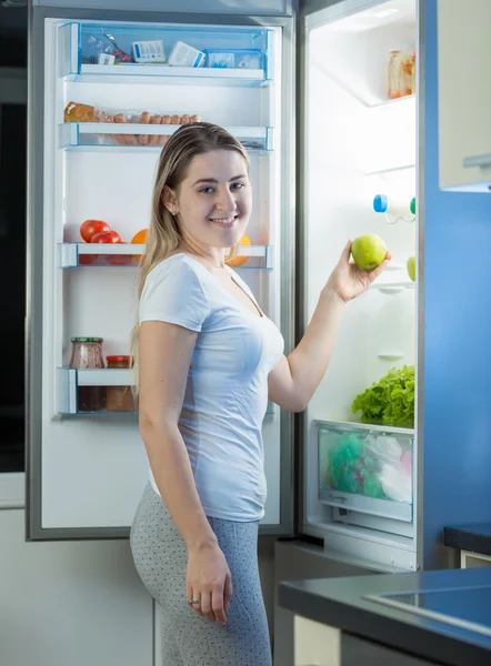 Hermosa mujer sonriente tomando manzana verde de la nevera por la noche —  Fotos de Stock