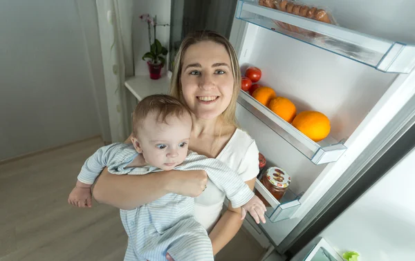 Улыбающаяся мать позирует с маленьким сыном рядом с открытым холодильником — стоковое фото