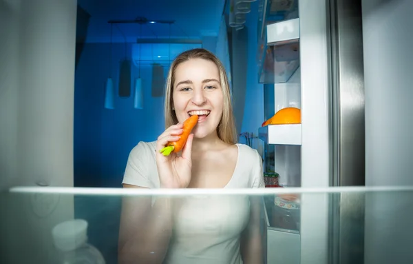 Gelukkig jonge vrouw nemen wortel uit koelkast en eten — Stockfoto