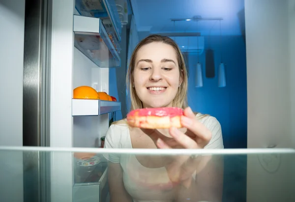 Mulher nova que abre o refrigerador e toma o grande donut à noite — Fotografia de Stock