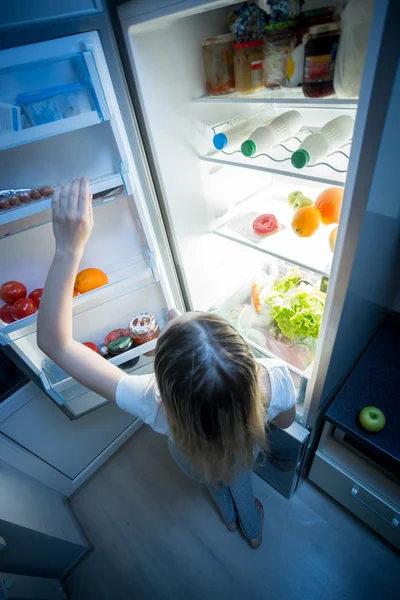 Young woman reaching for refrigerator top shelf at night — Stock Photo, Image