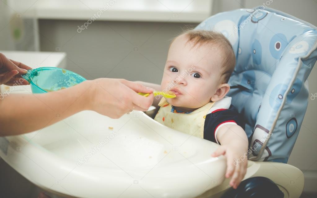Portrait of boy sitting in highchair at kitchen and eating from 