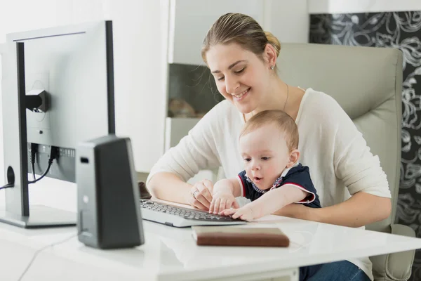 Mulher sorridente bonita trabalhando no computador com seu filho bebê — Fotografia de Stock