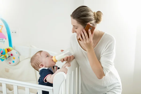 Retrato de la madre hablando por teléfono y alimentando a su bebé desde bo — Foto de Stock