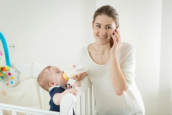 Retrato de una mujer sonriente hablando por teléfono mientras alimenta su bab —  Fotos de Stock