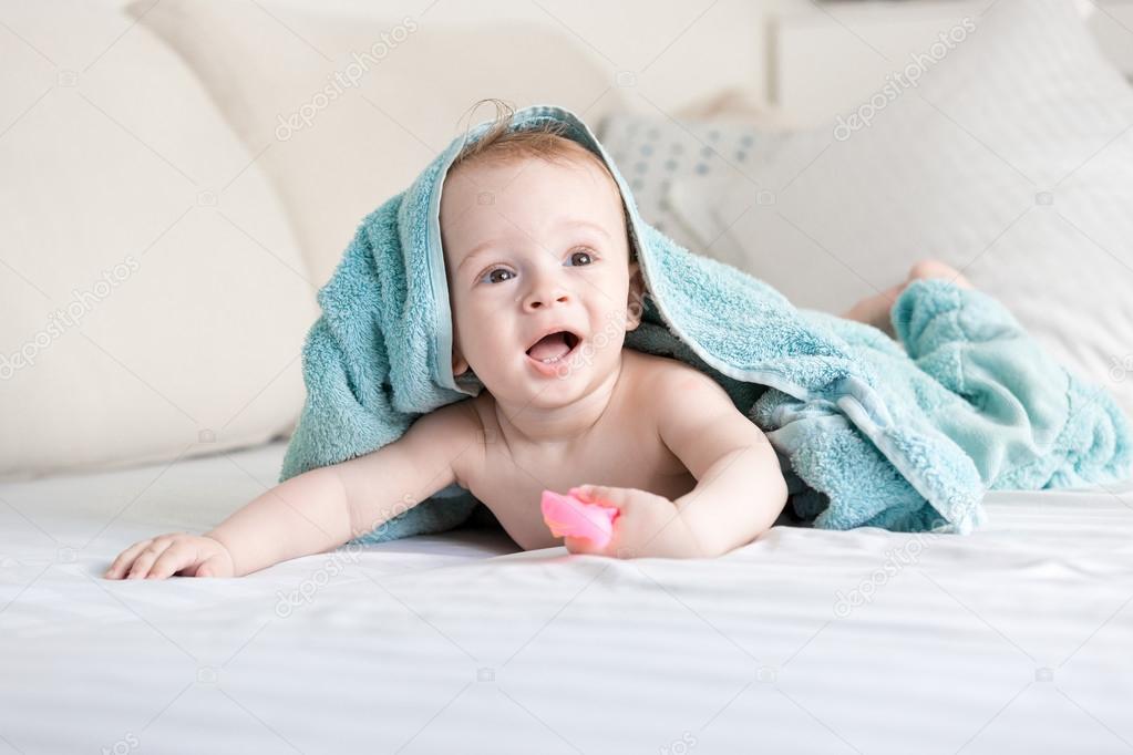 Happy smiling baby under blue towel crawling on bed with white s