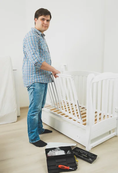 Handsome man in jeans and shirt assembling baby's cot — Stock Photo, Image