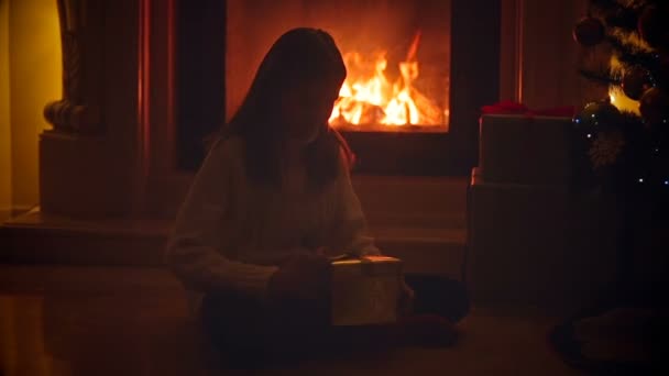 Симпатичная девушка сидит в темной гостиной у горящего камина и открывает коробку с подарком на Рождество — стоковое видео