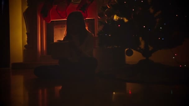 Dolly shot of happy girl sitting on floor at fireplace and opening box with Christmas gift — Stock Video