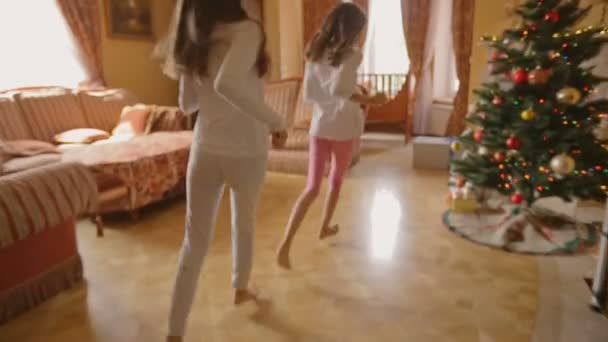 Две сестры в пижаме бегают в гостиной, чтобы открыть рождественские подарки — стоковое видео