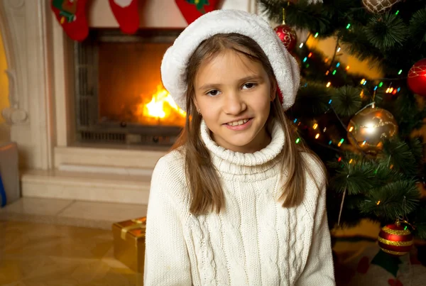 Счастливая улыбающаяся девушка в шляпе Санта сидит у горящего камина — стоковое фото