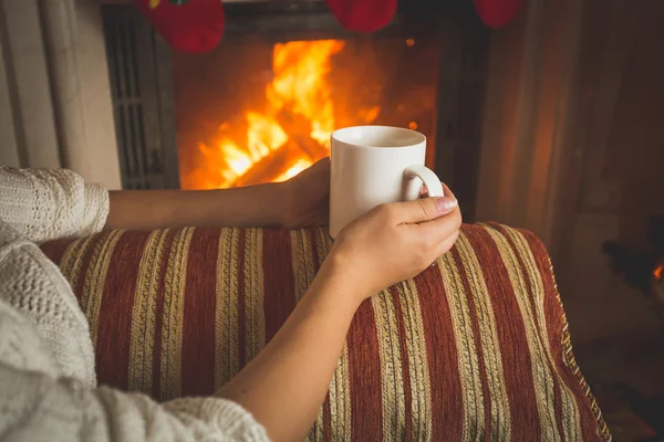 Toned image of woman sitting on sofa at fireplace and holding cu — Stock Photo, Image