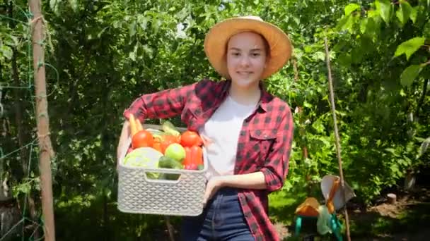 Усміхнена дівчина в капелюсі йде в полон з врожаєм у великій коробці. Свіжі, стиглі органічні овочі в коробці. Концепція малого бізнесу та вирощування органічних овочів на подвір'ї саду — стокове відео