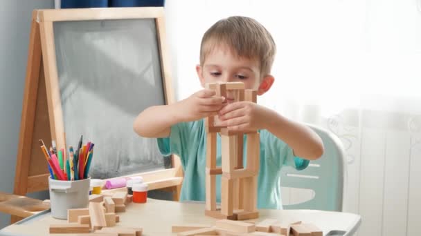 Menino sorridente feliz olhando para brinquedo blocos de madeira torre que ele construiu na mesa na sala de aula da escola. Conceito de crianças inteligentes e educação em casa durante o confinamento e auto-isolamento. — Vídeo de Stock