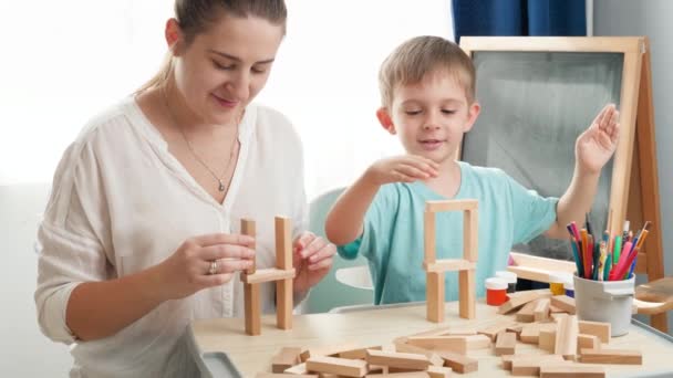 Медленное видео, как молодая мать с маленьким сыном играет и строит высокую башню из деревянных блоков или брикетов. Мать воспитывает и обучает своего ребенка дома во время изоляции и самоизоляции — стоковое видео