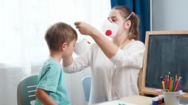 Ders çalışmadan ve okulda eğitim görmeden önce küçük çocuğa koruyucu maske takan genç bir kadın. Tecrit ve tecrit süresince çocukları virüsten korumak. Uzaktan eğitim ve eğitim...