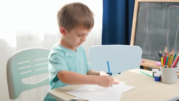 Koncentrált kisfiú rajzol vagy ceruzával ír, miközben otthon vagy az iskola osztálytermében ül az íróasztal mögött. Fogalom az oktatás és a távoli iskola otthon zárlat alatt és az önálló elszigetelés — Stock videók