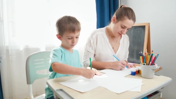 Χαμογελαστή μητέρα με μικρό γιο ζωγραφίζει με μολύβι. Παιδί κάθεται πίσω από το γραφείο του σχολείου και κάνει τα μαθήματά του με τους γονείς. Εκπαίδευση και απομακρυσμένο σχολείο στο σπίτι κατά τη διάρκεια αποκλεισμού και αυτο-απομόνωσης. — Αρχείο Βίντεο
