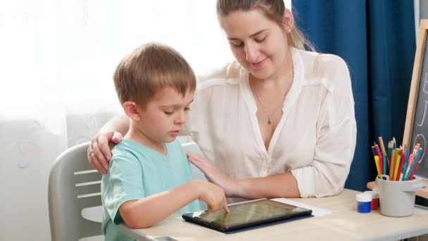 笑着妈妈拥抱和爱抚她在平板电脑上学习的聪明孩子。小男孩在家里的数字平板电脑上做作业 — 图库视频影像