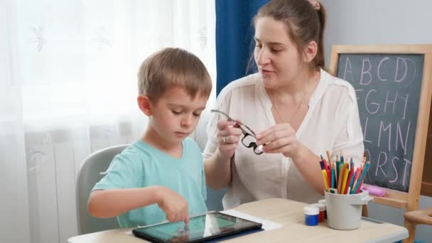 Mãe colocando óculos em seu filho inteligente brincando no computador tablet e fazendo lição de casa. Problema infantil com olhos e visão. — Vídeo de Stock