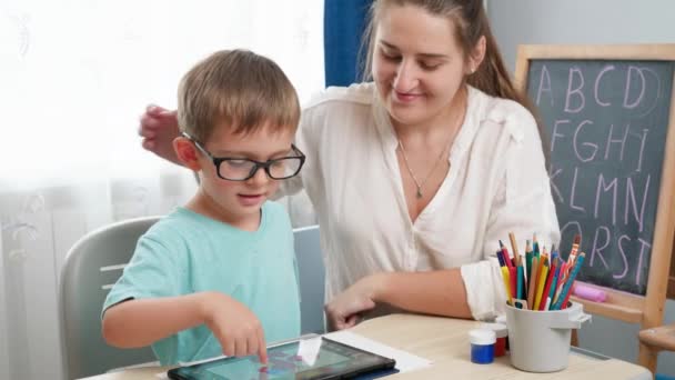 Gözlüklü çocuk tablet bilgisayarda oynuyor. Anne küçük oğluna sarılıp ödev yaparken gadet kullanıyor. Çocukların görme ve görme sorunları var. — Stok video