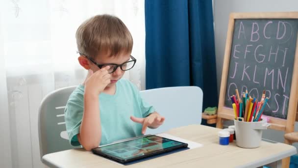 Маленький умный мальчик в очках изучает и использует цифровой планшетный компьютер. Обучение и обучение детей на дому с использованием гаджетов и современных технологий — стоковое видео