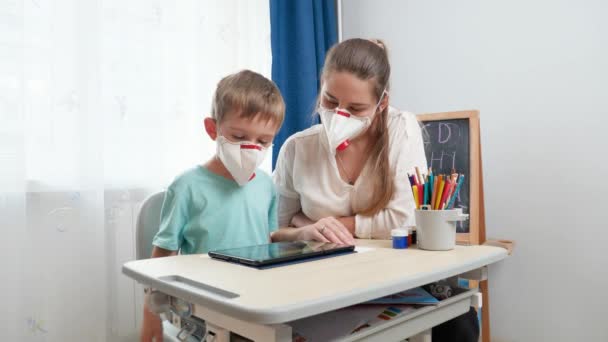 Matka s malým synem nosí ochranné masky lékařských respirátorů pomocí tabletu počítače. Dělat domácí úkoly a učit se doma během uzamčení a sebeizolace. Koncept vzdálené školy — Stock video