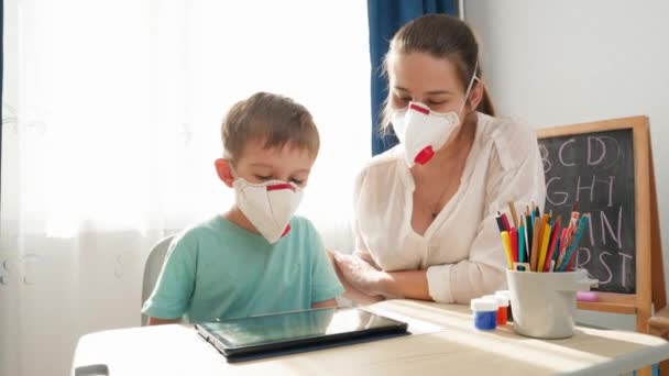 Nő tanít kisfiúnak táblagépet használni orvosi maszk lélegeztetővel. Távolsági oktatás és távoli iskola otthon a lezárás és az önelkülönítés idején. — Stock videók