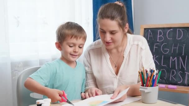 Mladá učitelka ukazuje své malé studentce, jak používat nůžky. Chlapec řezání papíru s nůžkami, zatímco domácí úkol. — Stock video