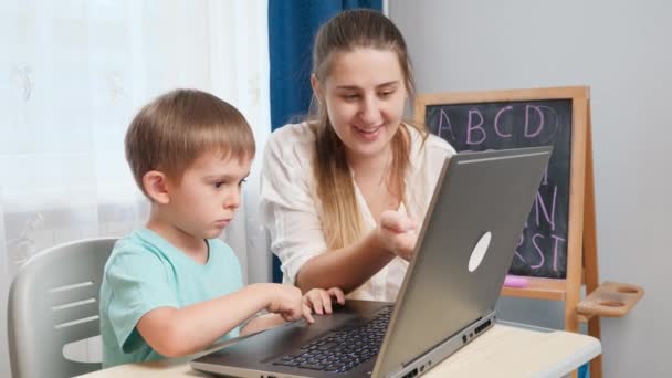 Улыбающаяся мать учит своего маленького сына пользоваться ноутбуком во время обучения и учебы дома. Концепция дистанционной школы и дистанционного образования на дому — стоковое видео