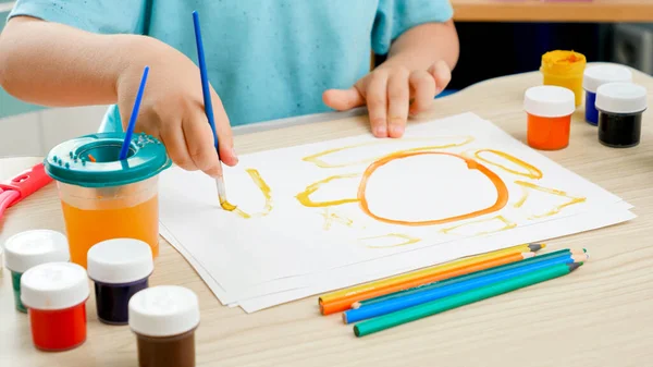 Ragazzino seduto dietro la scrivania e disegnare quadro colorato con vernice e pennelli. Educazione dei bambini a casa durante l'autoisolamento e l'isolamento. Concetto di arte e creatività dei bambini — Foto Stock