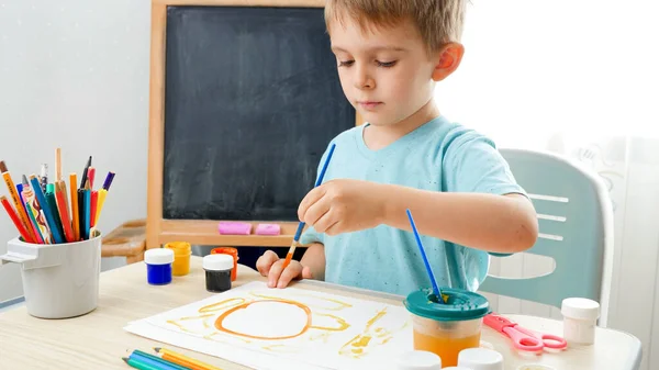 Портрет милого мальчика, заливающего кисть в красочную краску и делающего красивую картину. Творческий ребенок занимается живописью. Образование на дому во время изоляции — стоковое фото