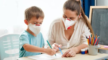 Küçük çocuk ve genç anne tıbbi maske takıyor ve evde ödev yapıyor. Tecrit ve tecrit süresince çocukları ve aileleri virüsten korumak. Uzak okul ve...