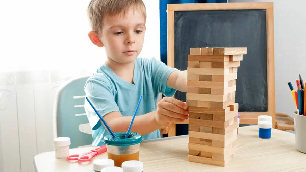 Умный мальчик вытаскивает деревянные блоки из высокой башни из кирпича. Концепция умных детей и домашнего образования во время изоляции и самоизоляции. — стоковое фото