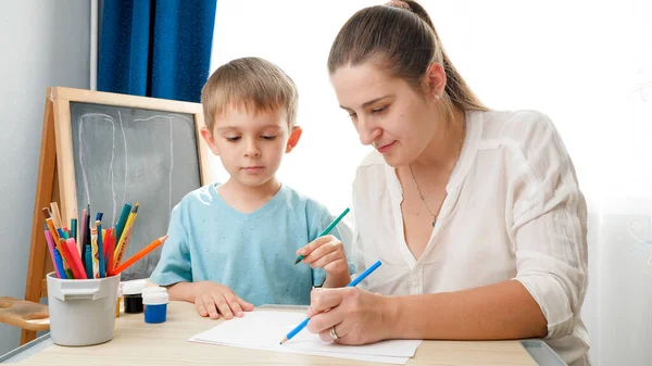 先生は学校の教室でカラフルな鉛筆で彼女の小さな幼児の息子を描きます。家庭での子育てと教育の概念。創造性と子供の芸術 — ストック写真