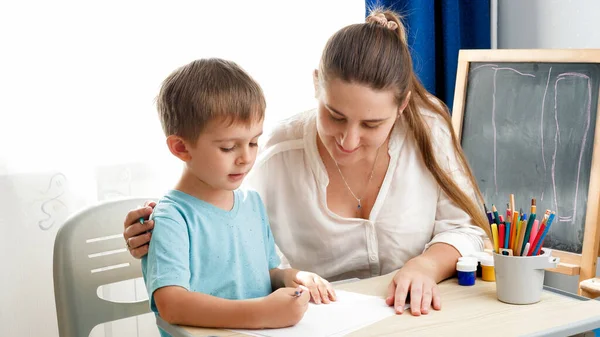 先生は学校の教室でカラフルな鉛筆で彼女の小さな幼児の息子を描きます。家庭での子育てと教育の概念。創造性と子供の芸術. — ストック写真