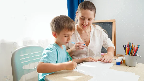 Leende mamma tittar på sin lille son rita eller skriva medan hon sitter bakom skrivbordet hemma klassrummet. Begreppsföräldrar och barn till utbildning och distansskola i hemmet under inlåsning och själv — Stockfoto