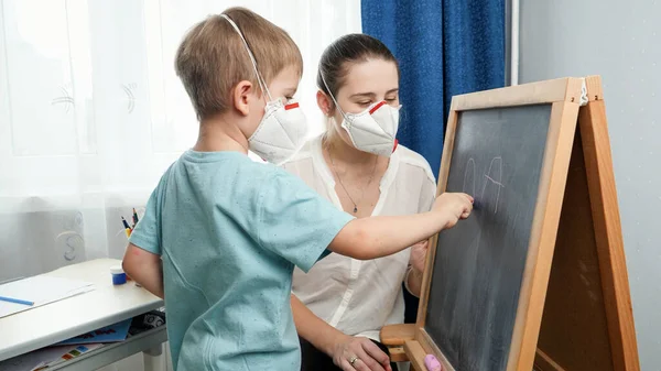 Petit garçon portant un masque médical protecteur écrivant des lettres sur un tableau noir avec de la craie. Éduquer et apprendre en classe sur un tableau noir pendant l'isolement et le confinement. École à distance à la maison — Photo